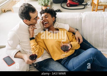 A happy multiracial couple relaxing on a sofa at home. This lifestyle image suits home life, relationship, and weekend moments, with subtle hints of wine and togetherness. Great for marketing materials about love, comfort, and casual entertaining. Stock Photo