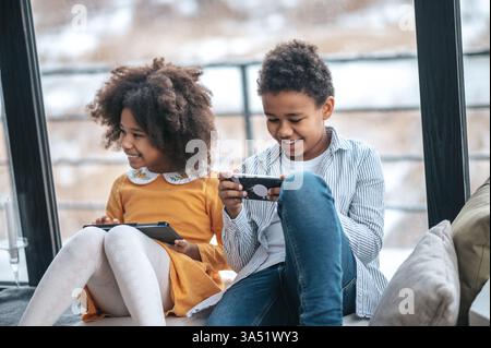 Two children indoors, spending time online on devices, capturing a modern digital family moment. Ideal for articles about kids and technology, screen time, and home life. Emphasizes youth, online learning, and connectivity. Stock Photo