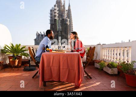 Multiracial couple on a rooftop balcony near Sagrada Familia in Barcelona, sharing a romantic aperitif with wine. Ideal for travel romance, couples photography, and urban lifestyle visuals. Stock Photo