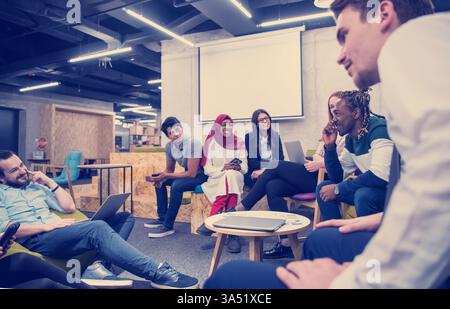 Smiling diverse startup team in a modern office, using laptops and a tablet during an informal meeting. The group includes colleagues from different backgrounds collaborating to share ideas and brainstorm strategies. Perfect for business, entrepreneurship and teamwork visuals. Stock Photo