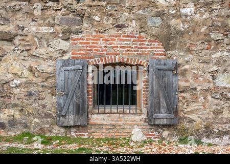 An aged, open wooden window featuring iron bars, set against a rugged backdrop of stone and brickwork. Stock Photo