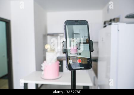 A pink cake is being photographed by a cell phone. The cake is placed on a table in a kitchen Stock Photo