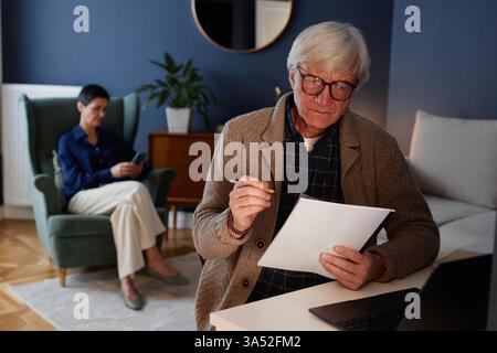 Elderly man studying document in living room while younger woman using cellphone in background creating a calm and focused atmosphere Stock Photo