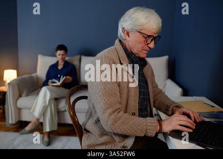 Senior man engaged in laptop work while sitting at desk, with woman reading on couch in background. Room with blue walls and cozy lighting creating a warm atmosphere Stock Photo