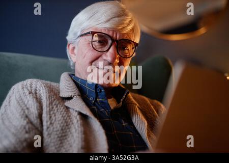 Elderly man with gray hair and glasses sitting on a cozy chair smiling while reading a book, creating a relaxed and comfortable ambiance in the room Stock Photo