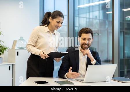 Two colleagues are collaborating on a project, reviewing information on a laptop in a bright, modern office setting. Stock Photo