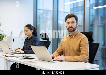 Two colleagues working side-by-side on laptops in a bright, modern office, focusing on their tasks. Stock Photo