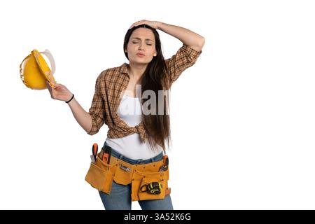 Tired female construction worker in a plaid shirt and tool belt holds her yellow helmet while touching her head, expressing exhaustion or stress, isol Stock Photo