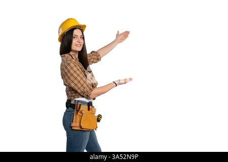 A female construction worker in a yellow hard hat and tool belt gestures with open hands, as if presenting something. She wears a plaid shirt and jean Stock Photo