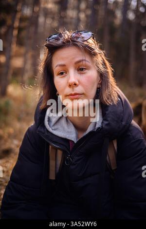 Close-up portrait of a young woman with sunglasses on her head, wearing a black jacket and hoodie, gazing into the camera with a calm expression in a Stock Photo