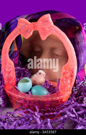Easter basket with wax child model head looking at new bird hatched from blue eggs lying on purple Easter grass. Mode is candle wax. Basket is vintage. Stock Photo