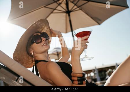 Side view, lying down with cocktail in hand. Beautiful young woman is at resort, vacation. Stock Photo
