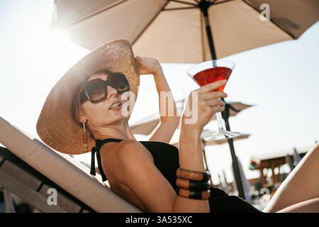 Side view, lying down with cocktail in hand. Beautiful young woman is at resort, vacation. Stock Photo