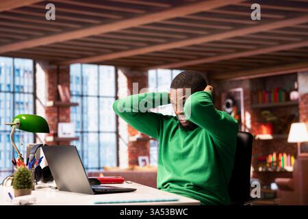 Teleworker in home office feeling hurting pain on the back of the head while working on laptop. Jaded remote employee suffering neck injury while solving tasks on notebook Stock Photo