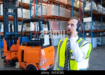 Using headset, warehouse manager coordinating logistics near forklift in warehouse, copy space Stock Photo