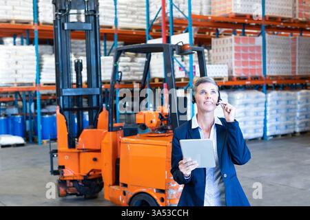 Coordinating logistics, warehouse manager using tablet and headset with forklift behind, copy space Stock Photo