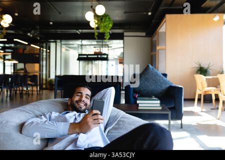 Relaxing in modern office, man smiling while using smartphone on bean bag Stock Photo