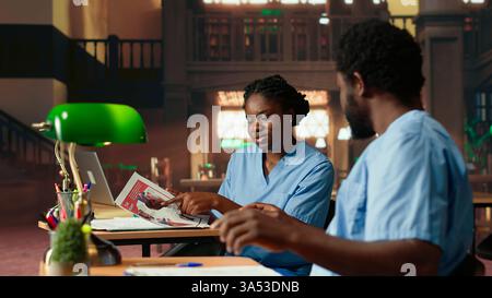 Future nurses studying anatomy class notes in a university library, reviewing courses and preparing for exams. Black med school students working together for their medical education. Camera B. Stock Photo