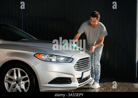 Guy is polishing the surface of automobile by using cloth. Man is with his car outdoors. Stock Photo