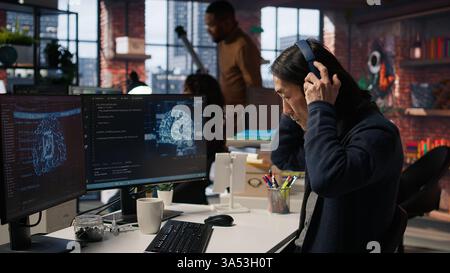 CTO manager in artificial intelligence focused startup drinking coffee, listening music and debugging applications. Asian technician putting headphones on, doing code testing, camera A Stock Photo