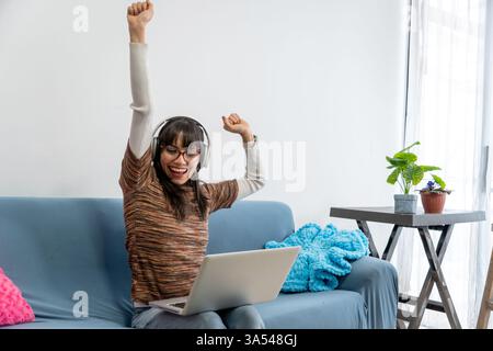 Happy businesswoman raising arms while using laptop and headphones, celebrating successful work from home Stock Photo
