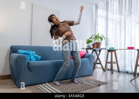 Carefree young woman enjoying music through headphones, dancing in her modern living room Stock Photo