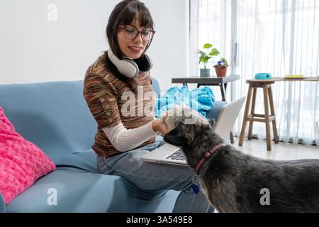 Young woman wearing headphones petting her dog while working from home using a laptop Stock Photo