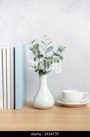 Modern home decor with eucalyptus branches in vase, books, and coffee cup on wooden desk against gray wall, creating cozy reading nook Stock Photo