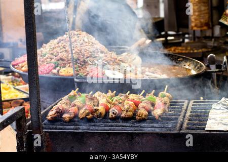 Meat and vegetable skewers grilling over smoky flames at a street food market, with a large paella pan in the background. Vibrant street food atmosphe Stock Photo