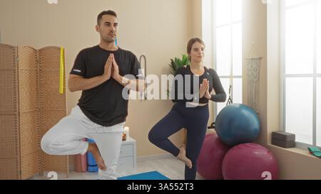 Man and woman practicing yoga in gym center performing tree pose on yoga mats next to exercise balls showing calm and focus in indoor environment. Stock Photo