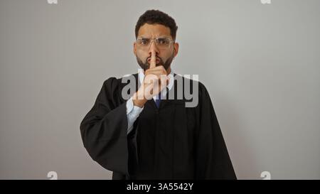 Young man with glasses in graduation gown making shushing gesture over white background, emphasizing silence Stock Photo