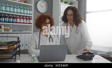 Women doctors collaborating in a hospital clinic room, reviewing patient data on a laptop, surrounded by medical equipment and healthcare documents in Stock Photo