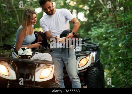 Holding yellow smartphone, showing something. Man and woman are on ATV outdoors. Stock Photo