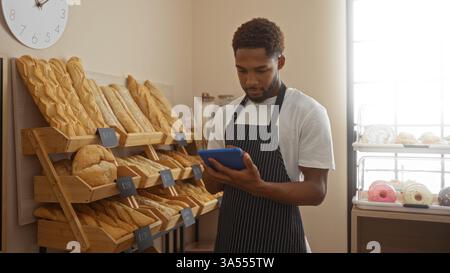 Young man working in bakery shop wearing apron and using tablet while standing next to various bread displays and a dessert shelf Stock Photo