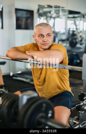 Fit mature man thoughtfully resting on barbell bench in gym setting Stock Photo