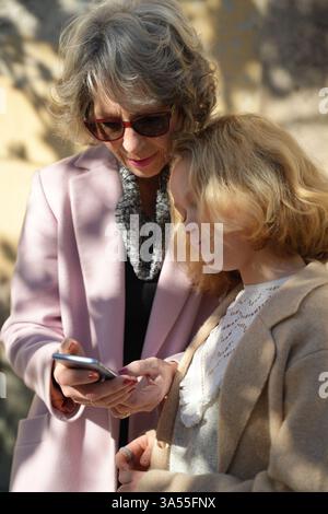 Grandmother in a pink coat and granddaughter enjoy looking at a smartphone together. Smiling and engaged, they experience bonding and learning through Stock Photo