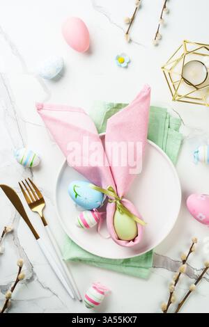 Easter table setting on white background with spring decorations. White plate, cutlery and colored eggs. Flat lay. Stock Photo