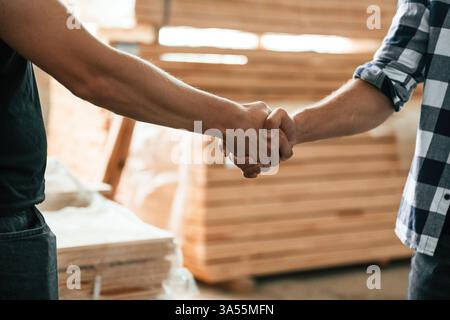 Close up view, doing handshake. Two workers are in the industrial wooden warehouse. Stock Photo