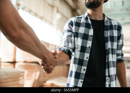 Close up view, doing handshake. Two workers are in the industrial wooden warehouse. Stock Photo