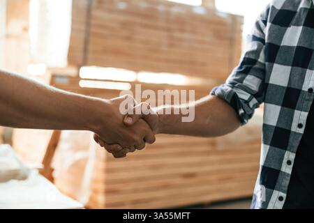 Close up view, doing handshake. Two workers are in the industrial wooden warehouse. Stock Photo
