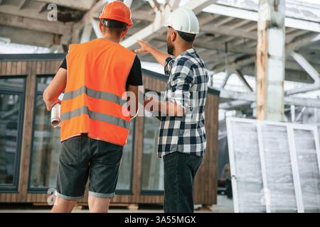 Builder is talking with manager or architect. Two workers are in the industrial wooden warehouse. Stock Photo