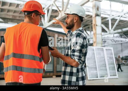 Builder is talking with manager or architect. Two workers are in the industrial wooden warehouse. Stock Photo