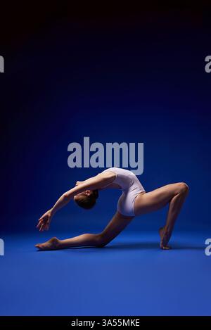 A female gymnast performing a deep backbend, showcasing incredible flexibility and strength. Elegant movement captured against a dark blue background. Stock Photo