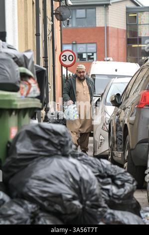 Avon Street, Birmingham 21st March 2025: A man struggles to take water to a mosque for Friday Prayers as he negotiates piles of rubbish and bin bags on Avon Street in the Sparkhill area of Birmingham as council refuse workers continue to strike on every working day until their demands are met. Talks on Thursday evening between the council and Unite failed to resolve issues. Residents have complained that one mountain of waste has been festering for several weeks outside their homes. More than 350 refuse workers began industrial action in January over the scrapping of the safety critical Waste Stock Photo