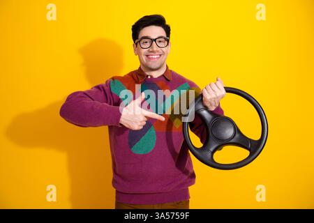 Young Man in Vibrant Jumper Smiling and Pointing at a Steering Wheel with a Bright Yellow Background Depicting a Fun Stock Photo