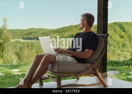 Young male freelancer seated on a wooden chair, with laptop, enjoying nature and a beautiful forest view while working remotely. Work-life balance Stock Photo