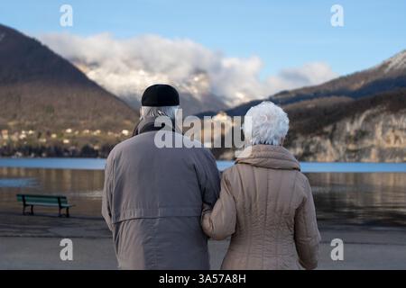 Elderly Couple Holding Hands, Facing Lake Annecy in Winter – Peaceful Retirement Moment in France Stock Photo
