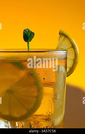 One glass of a ice tea with lime and lemon slices in it. The drink is served in a glass with mint leaves on the top. Warm orange background. Stock Photo