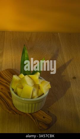 A bowl of pineapple slices is on a wooden cutting board. The bowl is white and the pineapple slices are yellow. The pineapple slices are cut Stock Photo