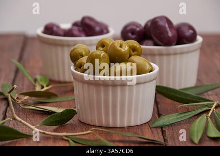 Black and green olives in round bowls, on a rustic wooden background with olive leaves Stock Photo